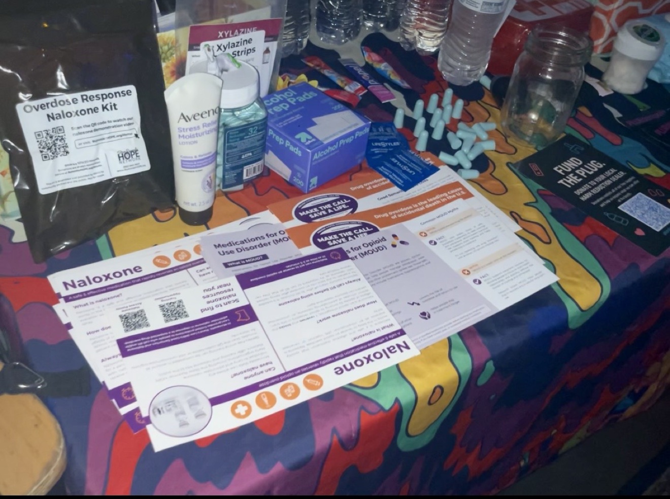 Close-up of harm reduction supplies on the booth table: naloxone kits, test strips, earplugs, and educational flyers